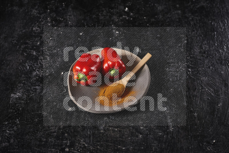 A wooden spoon full of ground paprika powder with red bell peppers, all on a pottery plate on black background