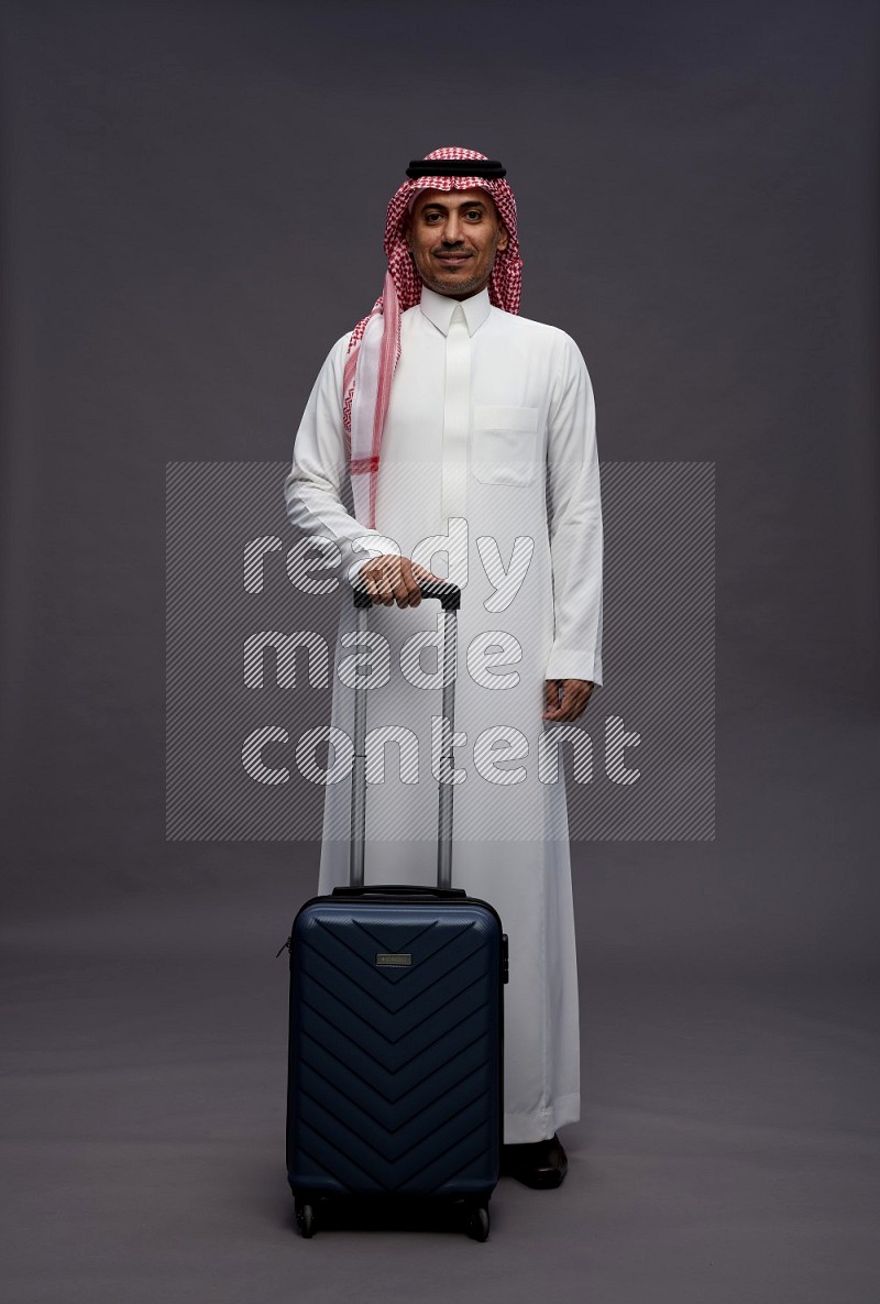 Saudi man wearing thob and shomag standing holding bag on gray background
