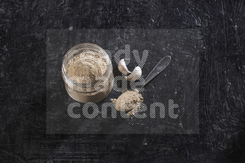 A glass jar full of garlic powder with a metal spoon full of the powder on a textured black flooring