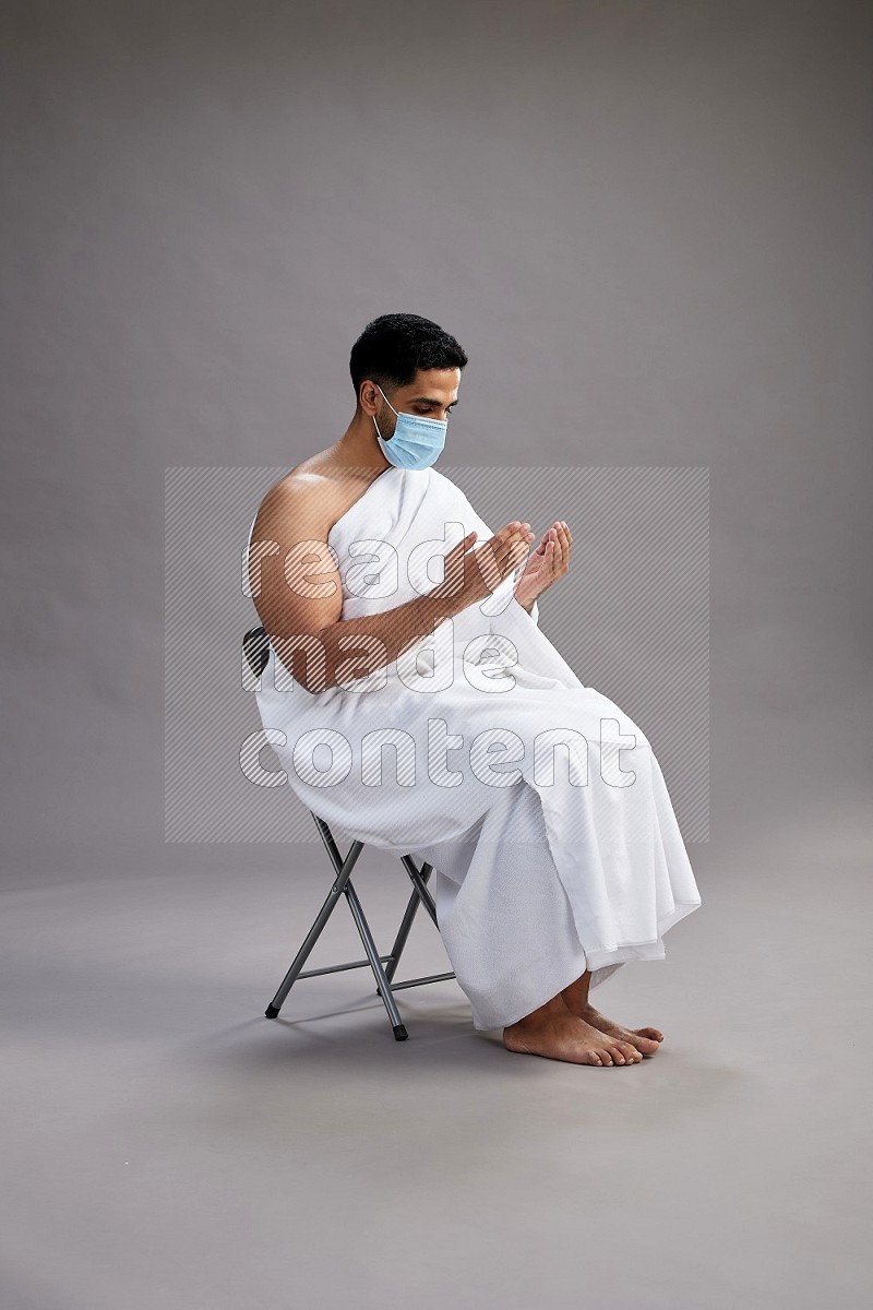 A man wearing Ehram with face mask sitting on chair dua'a on gray background