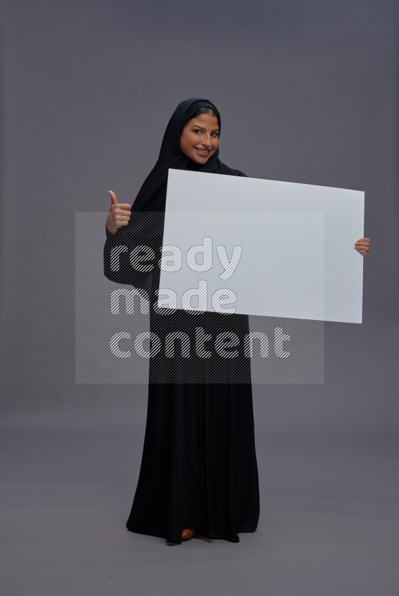 Saudi woman wearing Abaya standing holding white board on gray background