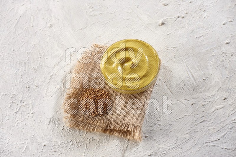 A glass bowl full of mustard paste set on a burlap piece with some mustard seeds on a textured white flooring in different angles