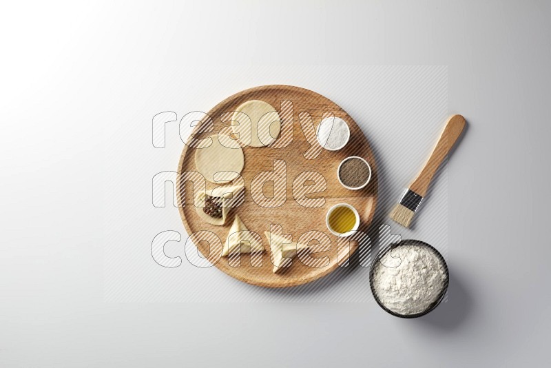 two closed sambosas and one open sambosa filled with meat while flour, salt, black pepper and oil with oil brush aside in a wooden dish on a white background