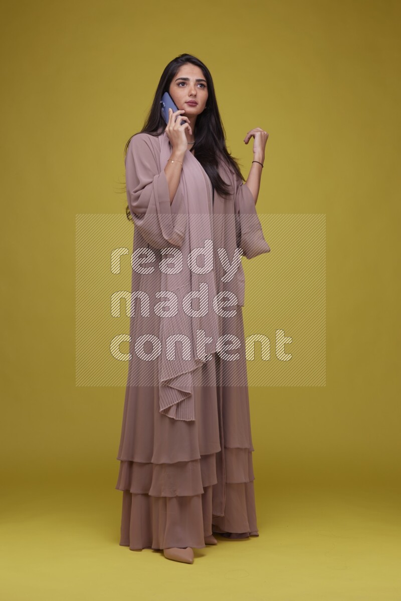 A woman Calling on a Yellow Background wearing Brown Abaya