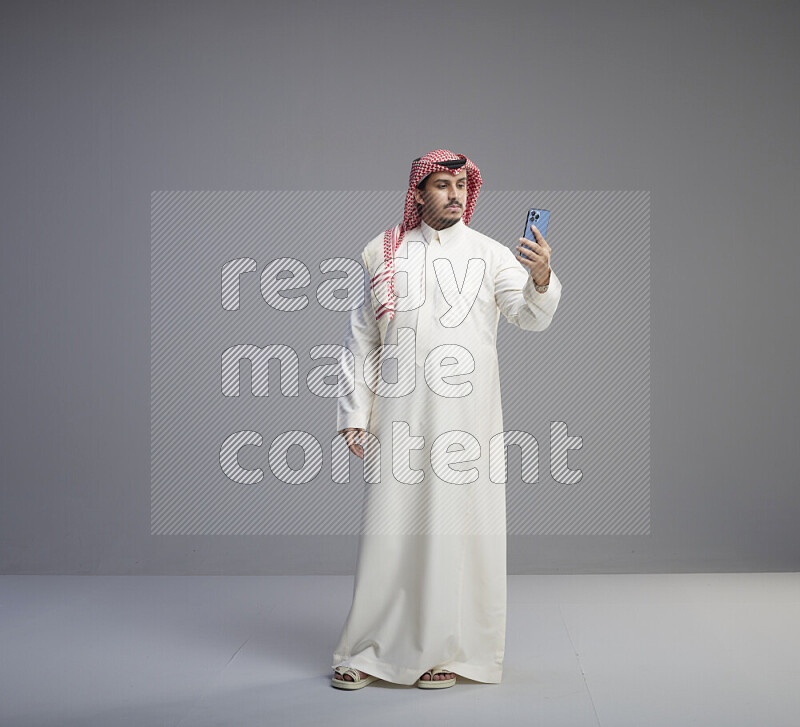 A Saudi man standing wearing thob and red shomag taking selfie on gray background