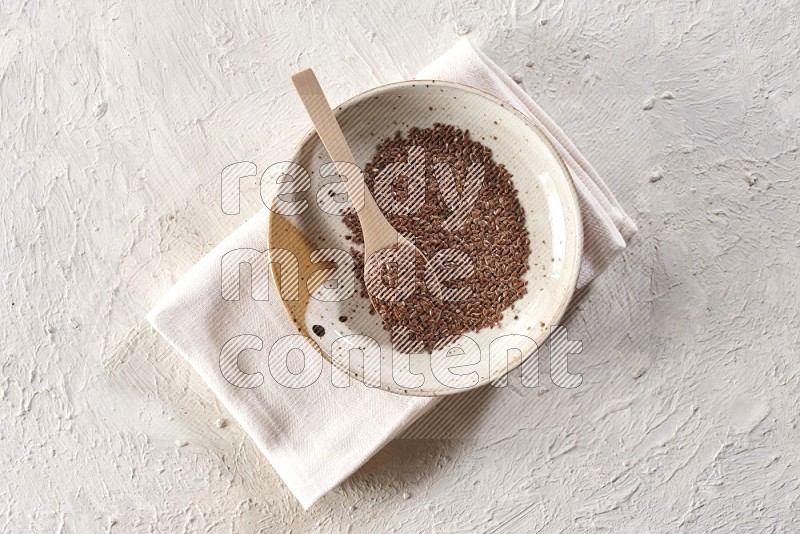 A multicolored pottery plate full of flax seeds with a wooden spoon full of the seeds on a napkin on a textured white flooring