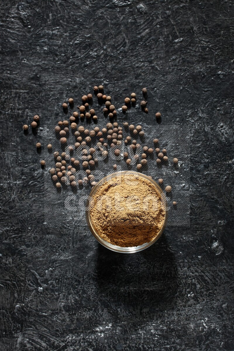 A glass bowl full of allspice powder and whole balls spreaded on a black flooring