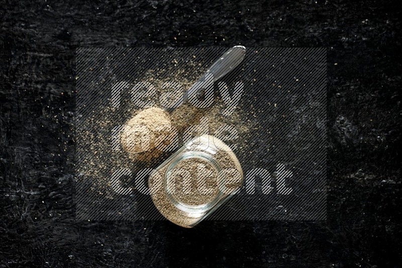 A glass spice jar and metal spoon full of cardamom powder on textured black flooring