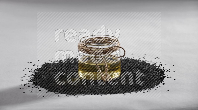 A glass jar full of black seeds oil surrounded by the seeds on a white flooring