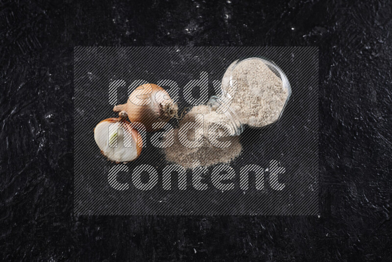 A glass jar full of onion powder flipped with some spilling powder on black background