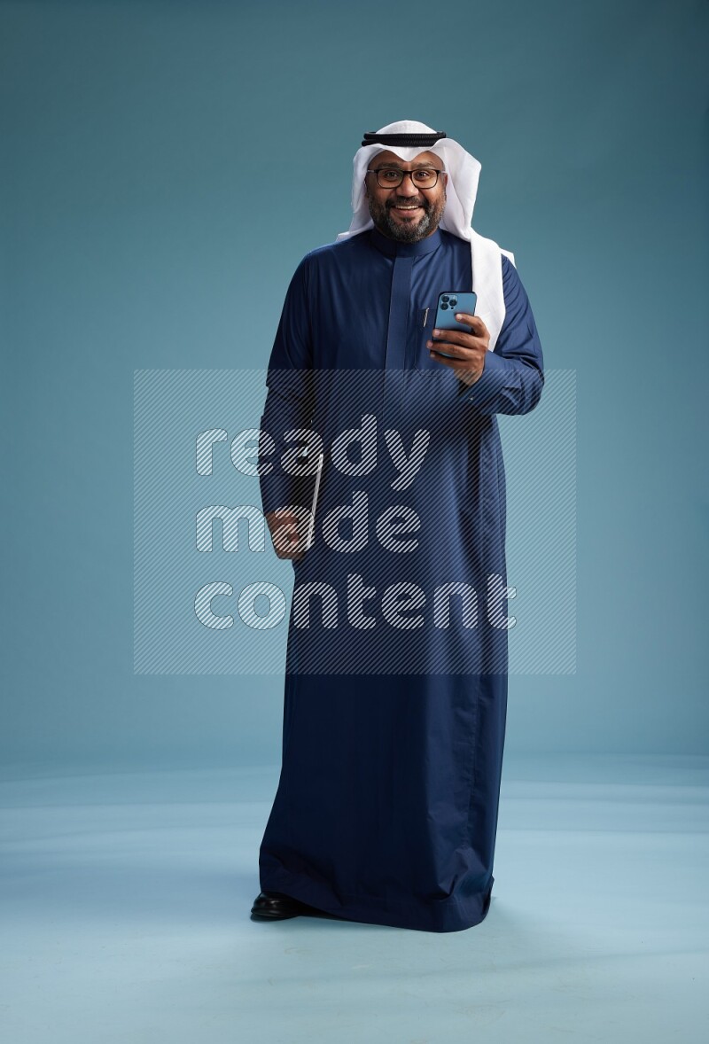 Saudi Man with shimag Standing texting on phone on blue background