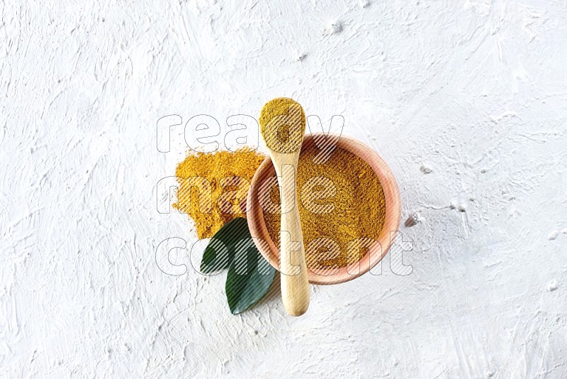 A wooden bowl and wooden spoon full of turmeric powder on textured white flooring