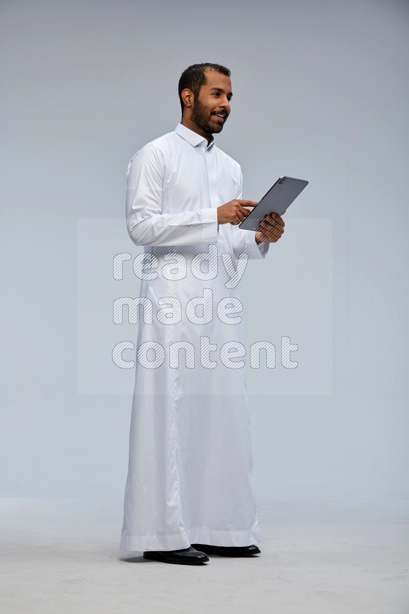 Saudi man wearing Thob standing working on tablet on Gray background