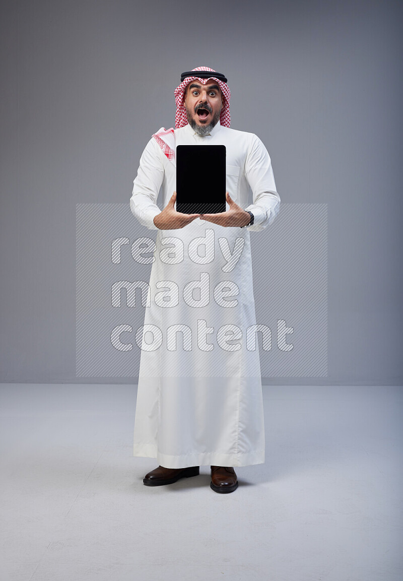 Saudi man Wearing Thob and red Shomag standing showing tablet to camera on Gray background