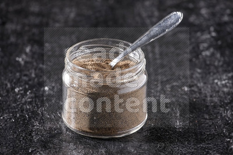 بودرة الفلفل الأسود في وعاء زجاجي مع ملعقة معدنية علي خلفية سوداء