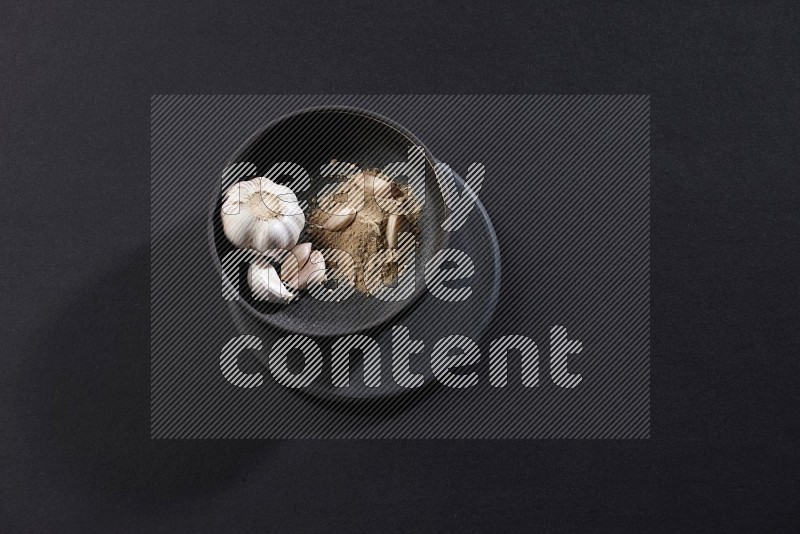 A black pottery bowl full of garlic powder, some cloves and a whole garlic bulb on a black flooring