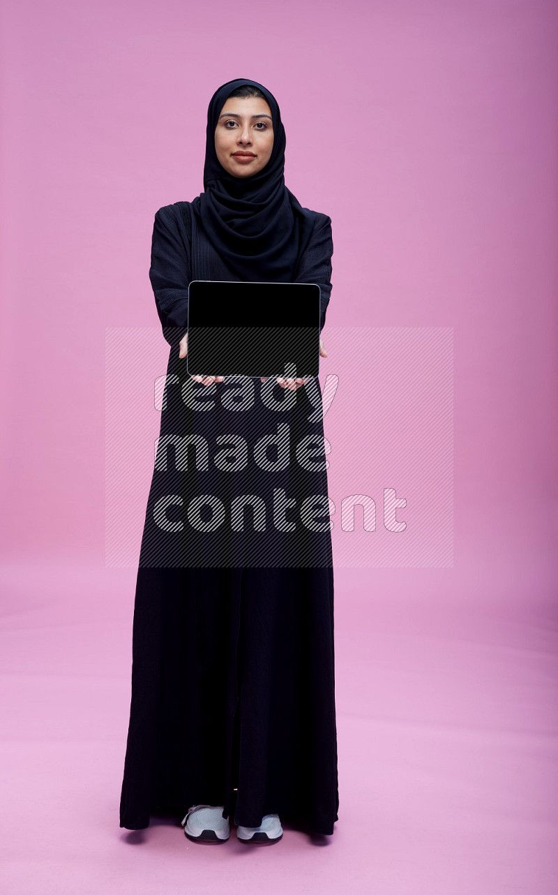 Saudi woman wearing Abaya standing showing tablet to camera on pink background