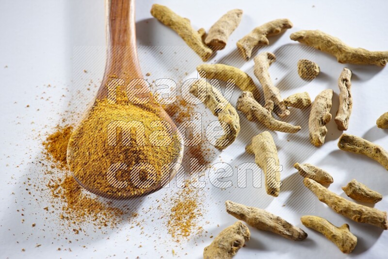 A wooden ladle full of turmeric powder and dried turmeric fingers beside it on white flooring