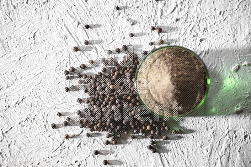 A glass cup full of black pepper powder and black pepper spread on a textured white flooring