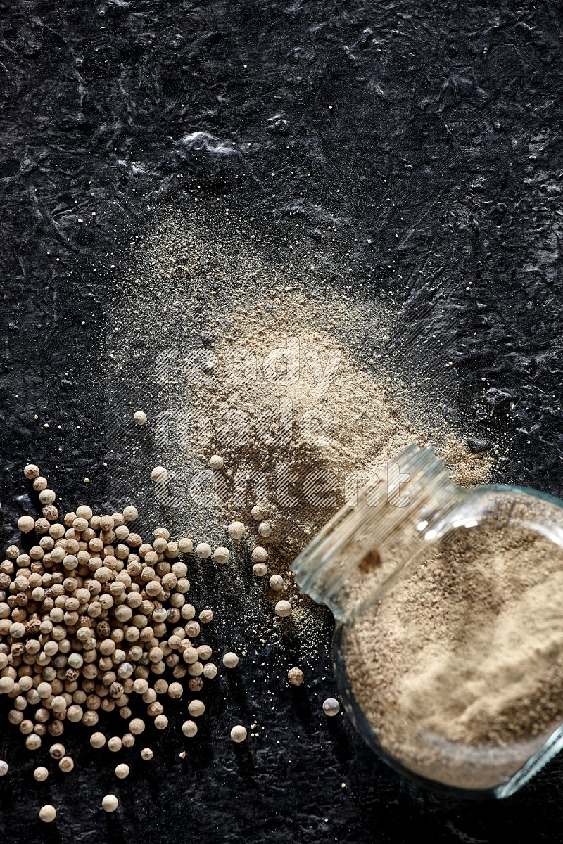 A flipped herbal glass jar full of white pepper powder with spilled powder and pepper beads on textured black flooring