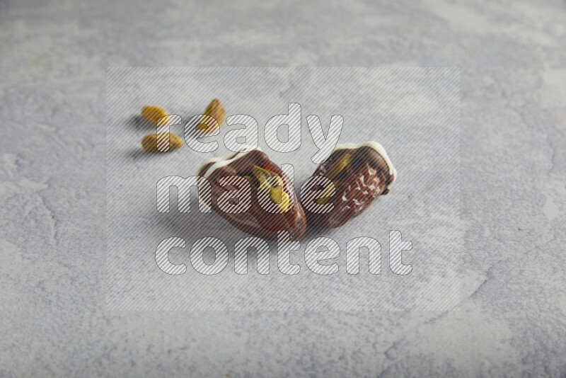 two pistachio stuffed dates covered with dark chocolate with white chocolate drizzle with unroasted pistachios on a light grey background