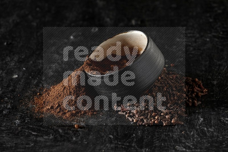 وعاء فخاري أسود ممتلئ ببودرة القرنفل والبودرة تناثرت منه مع الحبوب على خلفية سوداء