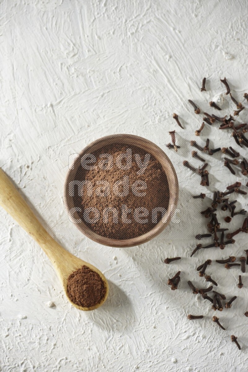 A wooden bowl and wooden spoon full of cloves powder with cloves spread on textured white flooring