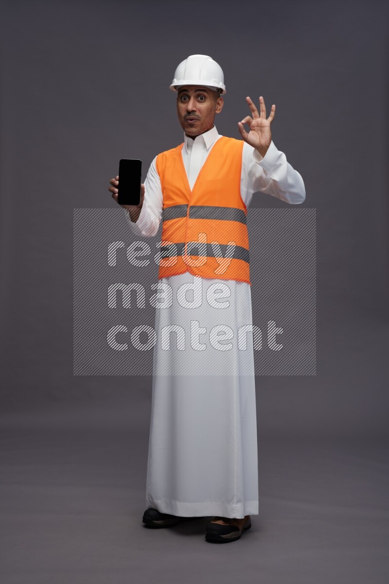Saudi man wearing thob with engineer vest standing showing phone to camera on gray background