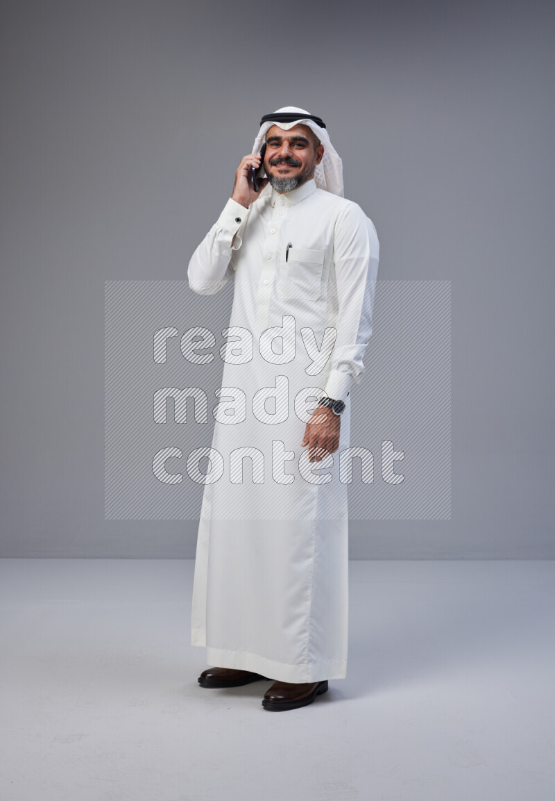 Saudi man Wearing Thob and white Shomag standing talking on phone on Gray background
