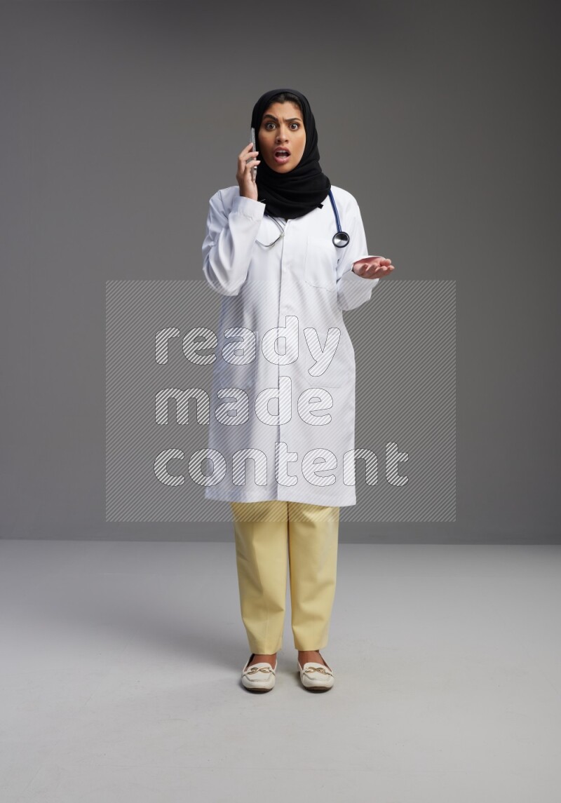 Saudi woman wearing lab coat with stethoscope standing talking on phone on Gray background