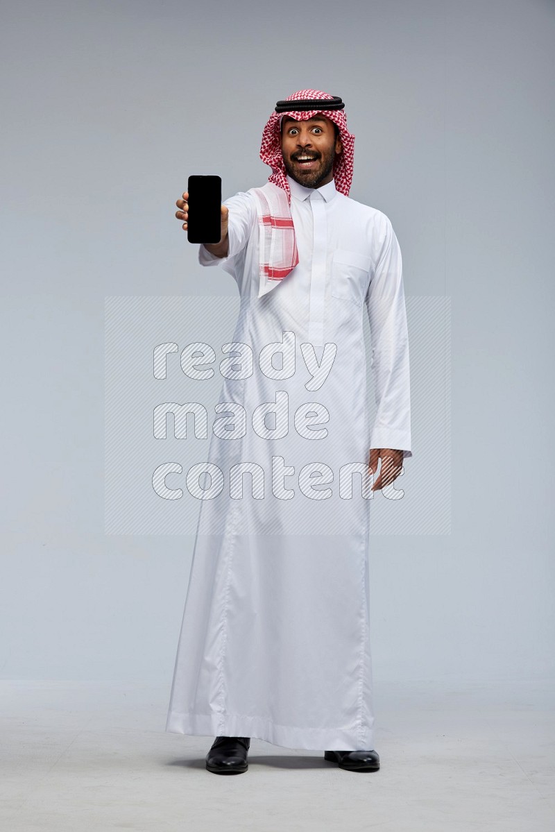 Saudi man Wearing Thob and shomag standing showing phone to camera on Gray background