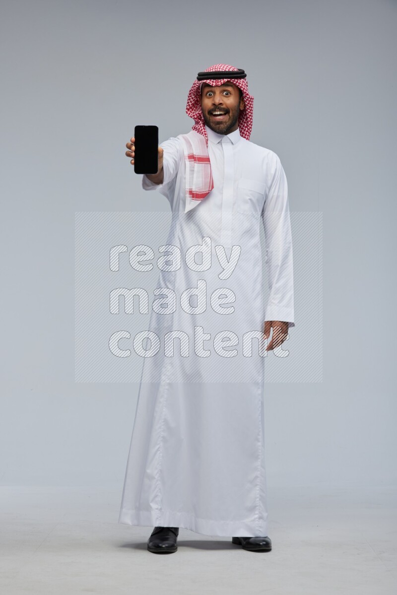 Saudi man Wearing Thob and shomag standing showing phone to camera on Gray background