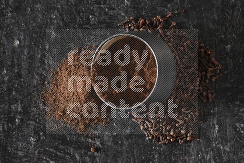 A black pottery bowl full of cloves powder spilled out of it with whole grains on a textured black flooring
