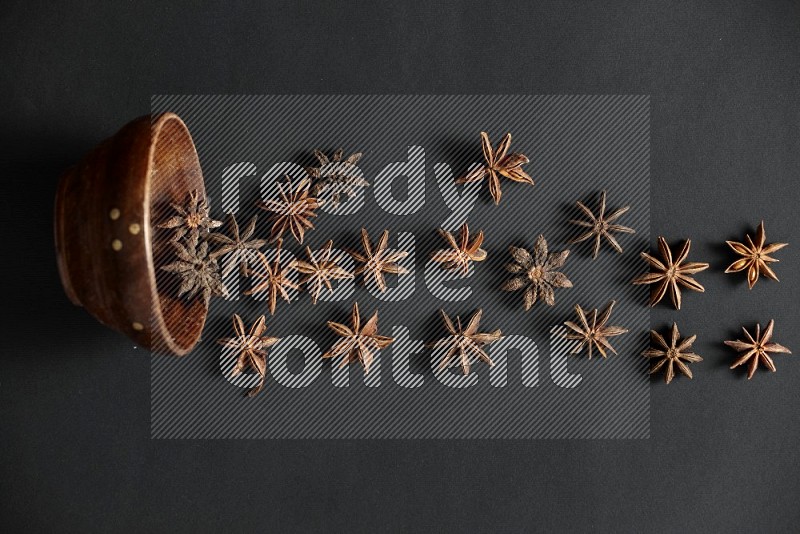 Star anise lined out of wooden bowl across the frame on black background