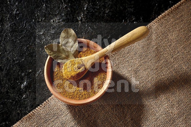 وعاء خشبي وملعقة خشبية ممتلئان ببودرة الكركم على قماش الخيش موضوعان على خلفية سوداء