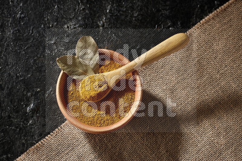 A wooden bowl and a wooden spoon full of turmeric powder on burlap fabric on textured black flooring