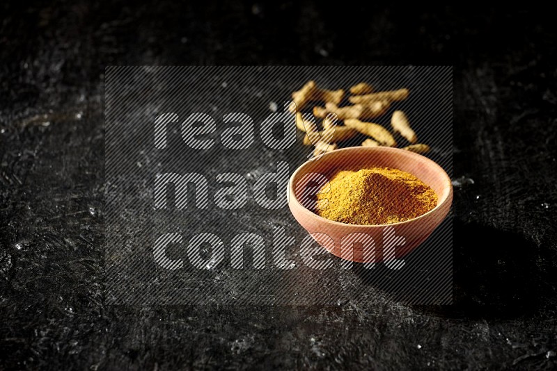 A wooden bowl full of turmeric powder with dried turmeric fingers on textured black flooring