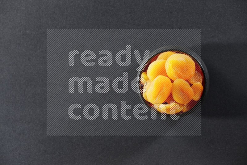 A black pottery bowl full of dried apricots on a black background in different angles
