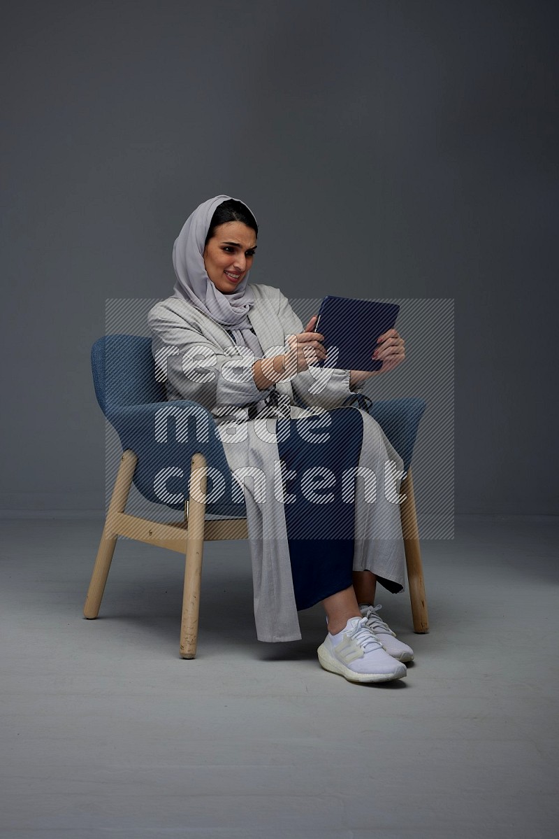 A Saudi female wearing a light gray Abaya and head scarf sitting on a dark grey chair while holding her iPad on a grey background