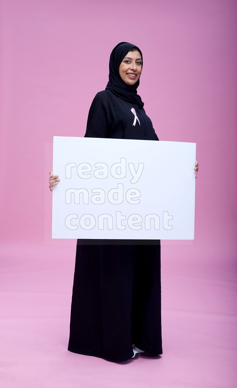 Saudi woman wearing pink ribbon on Abaya standing holding board on pink background