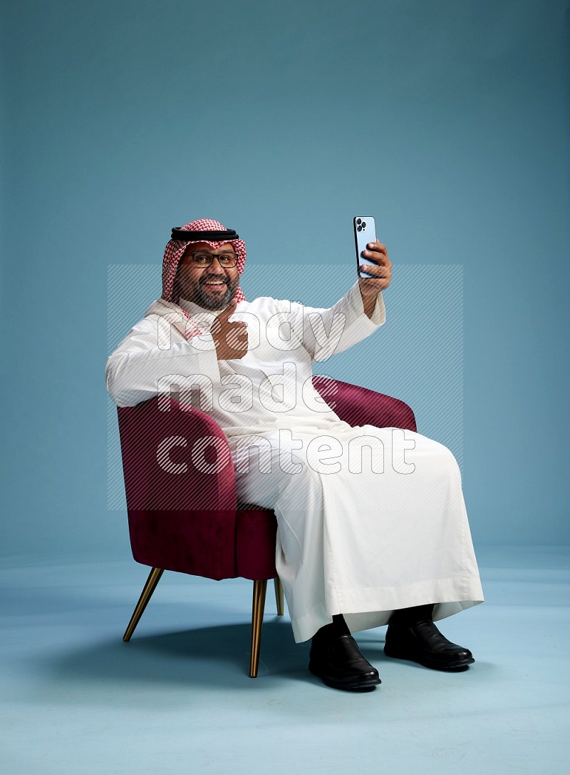Saudi Man with shimag sitting on chair taking selfie on blue background