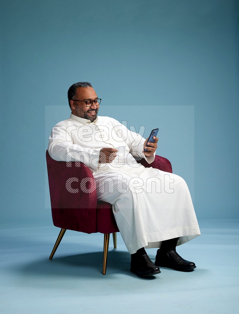 Saudi Man without shimag sitting on chair holding ATM while talking on phone on blue background