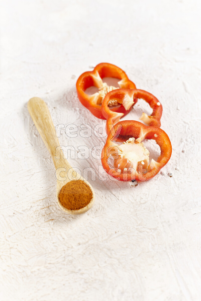 A wooden spoon full of ground paprika powder with red bell pepper slices beside it on white background