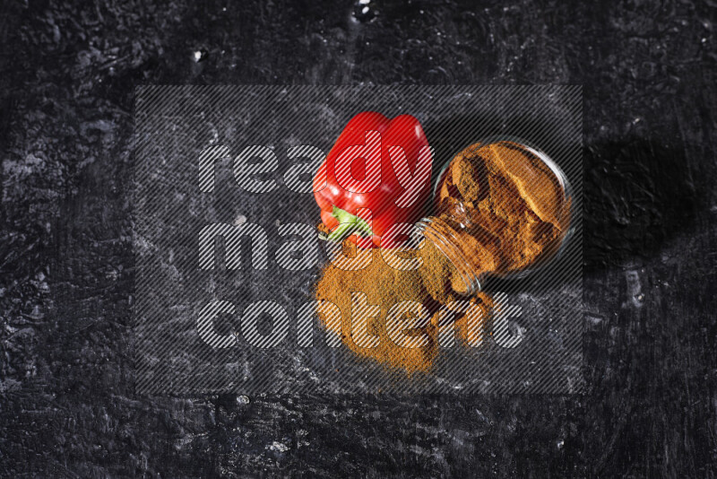 A glass jar full of ground paprika powder flipped with some spilling powder on black background