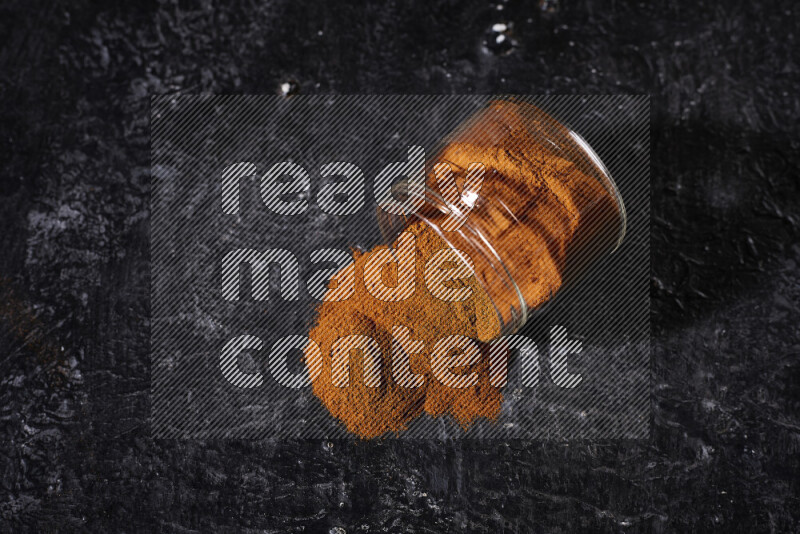 A glass jar full of ground paprika powder flipped with some spilling powder on black background