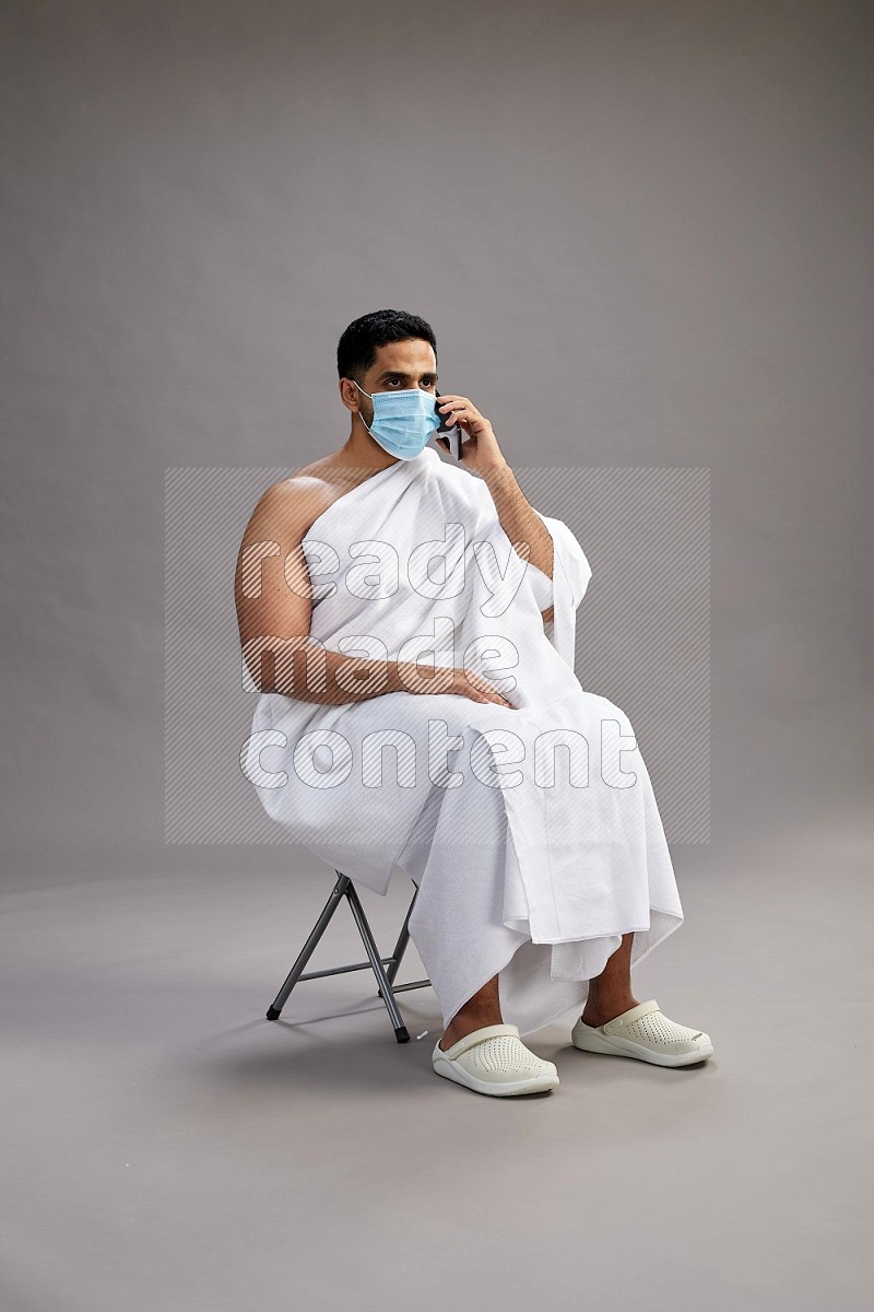 A man wearing Ehram with face mask sitting on chair talking on phone on gray background