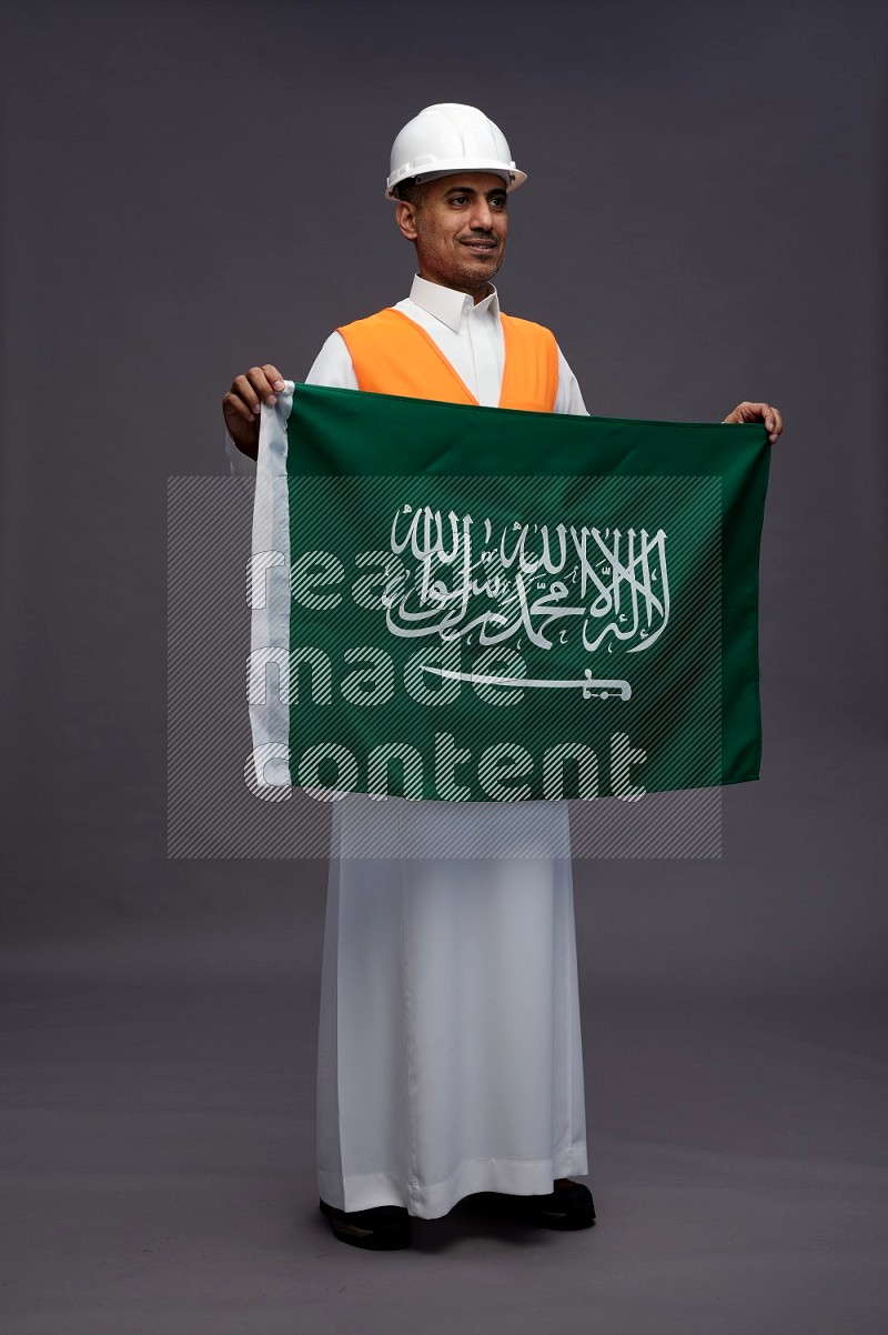 Saudi man wearing thob with engineer vest standing holding Saudi flag on gray background