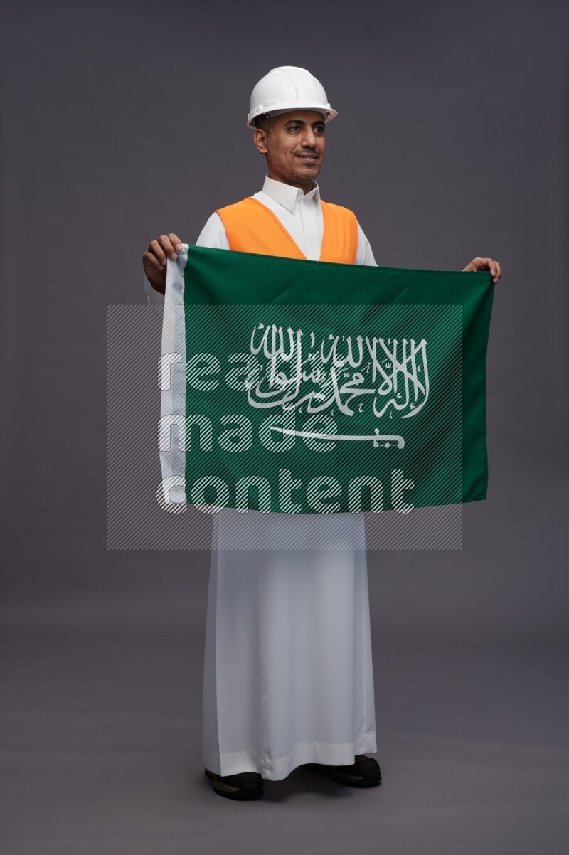 Saudi man wearing thob with engineer vest standing holding Saudi flag on gray background