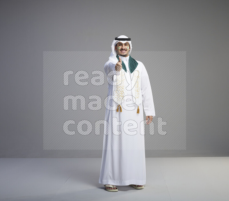 A Saudi man standing wearing thob and white shomag with face painting and Saudi flag scarf on gray background