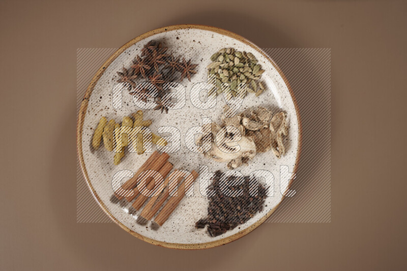 An assorted whole spices on a flat beige plate including fresh and dried ginger, cinnamon sticks, cardamom, turmeric fingers, cloves, star anise on a beige background